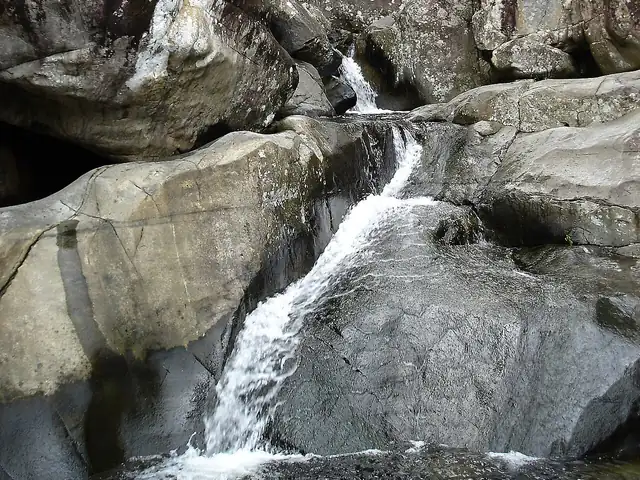 Water pours in a white cascade between large, smooth grey granite boulders into a rock pool.