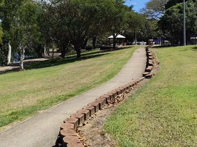 A concrete footpath edged with a low brick border winds up a grassy slope towards shady trees and a picnic shelter.