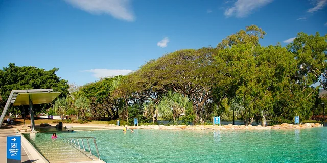A wide, clear lagoon pool edged with boulders and shady trees, with a ramp and handrail leading into the water and an access sign beside it.
