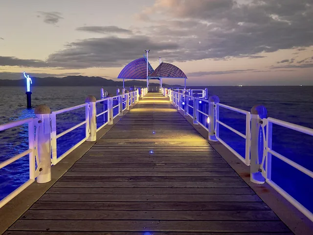 A long timber pier lit with blue lights at dusk, with white railings on both sides and a curved shade roof at the far end, over the sea.