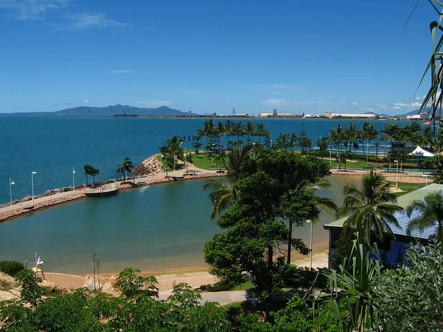 Seen from above, a large enclosed sea-water pool curves along the shore, edged by a rock wall, sand and palm trees, with the sea and distant hills beyond.